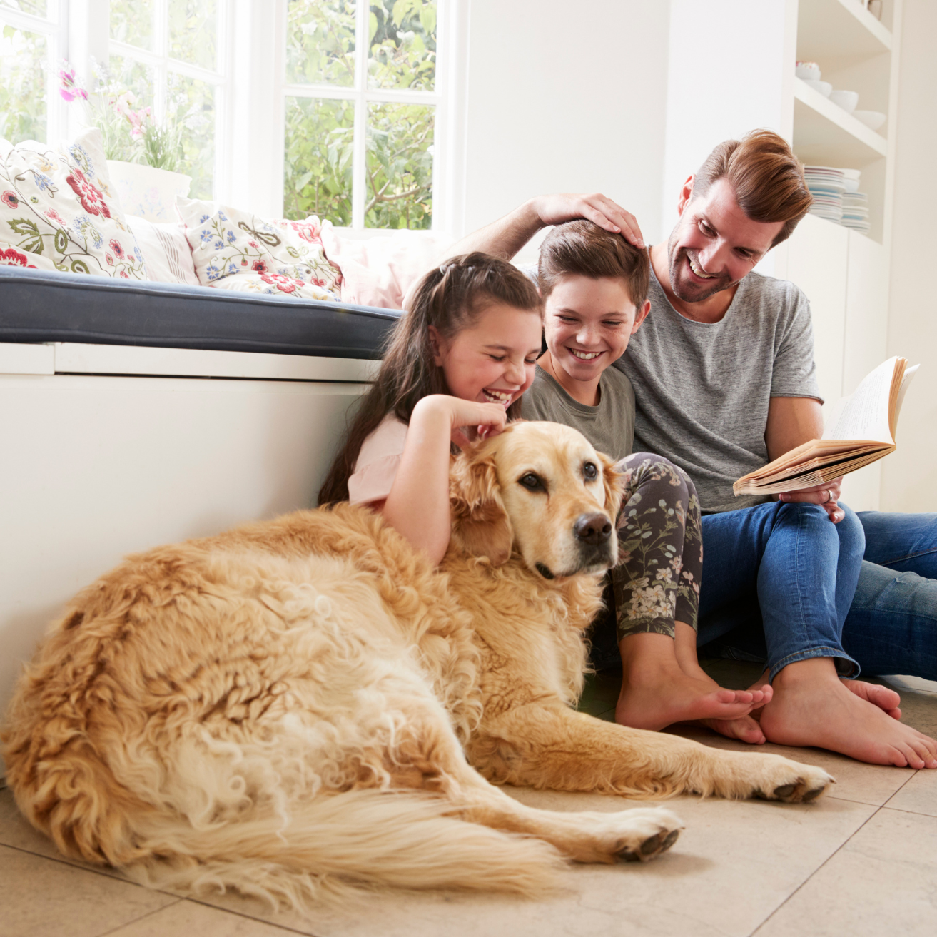 Eine Familie sitzt auf dem Boden, der Vater hat ein Buch in der Hand. Zusammen mit der Familie liegt noch ein Hund auf dem Boden.